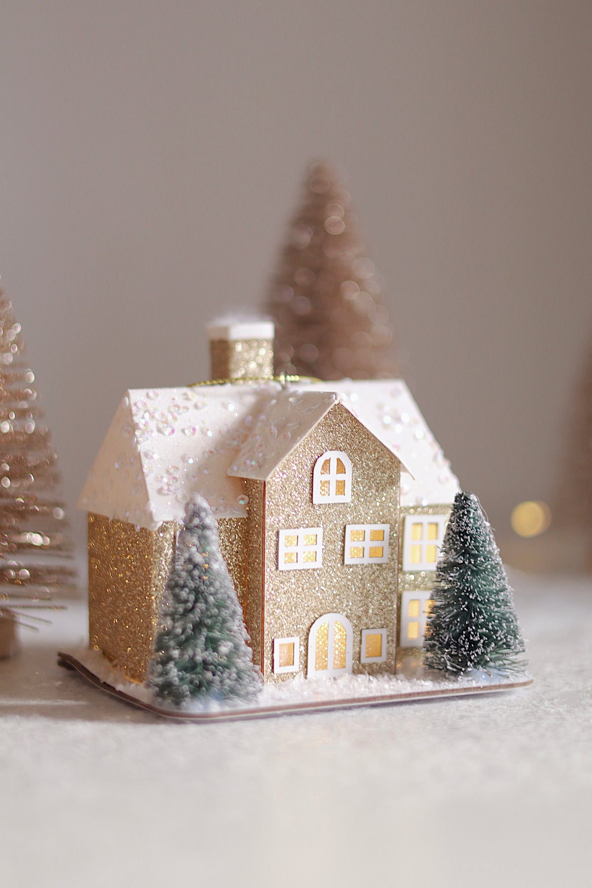A miniature gold glitter Christmas house ornament with a snowy roof, glowing yellow windows, and two small frosted green trees, photographed on a soft neutral background.