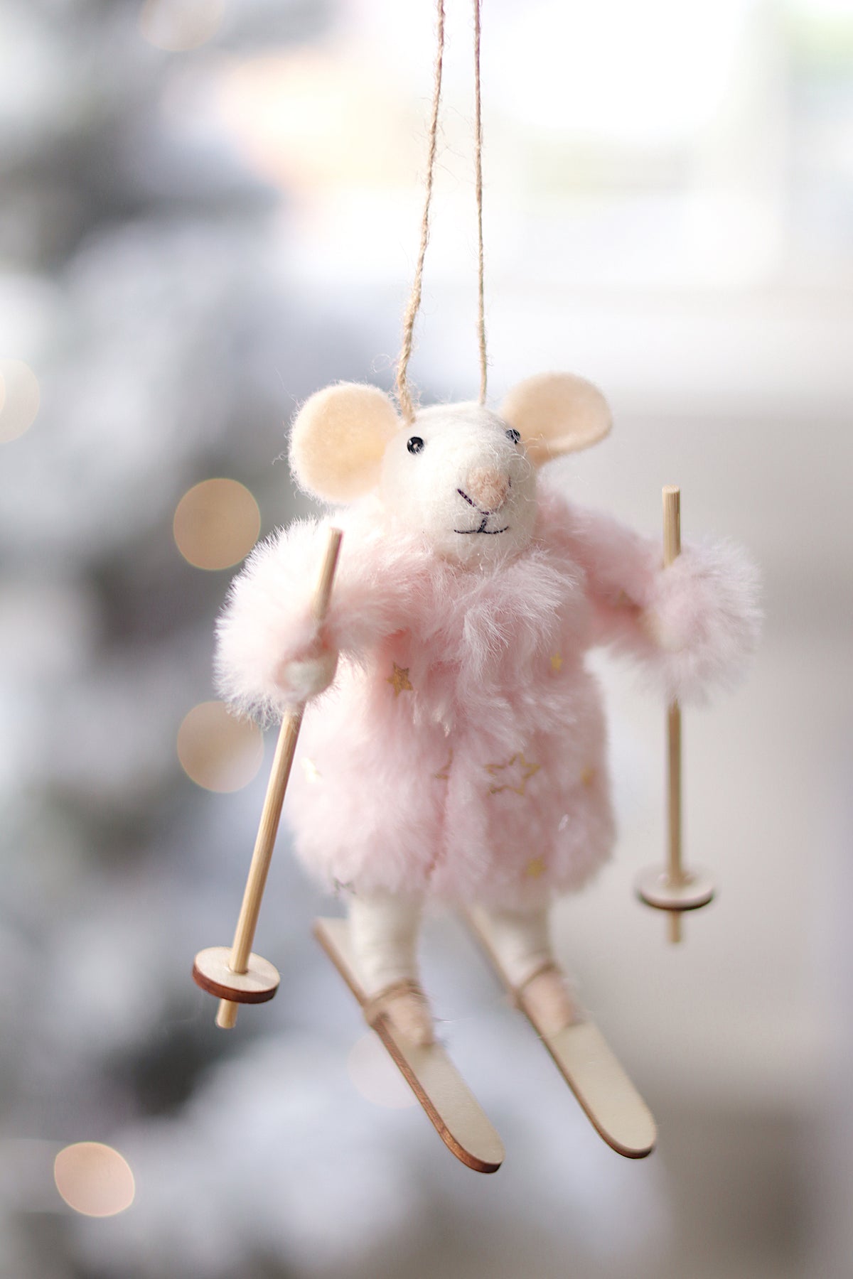 A plush hanging ornament shaped like a small white animal wearing a fluffy pink coat and holding wooden ski poles and skis, photographed against a softly blurred light background.